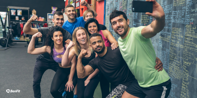 Gruppe von glücklichen Fitness-Enthusiasten posiert für ein Selfie im Fitnessstudio vor einer Tafel mit Trainingsübungen und Notizen.