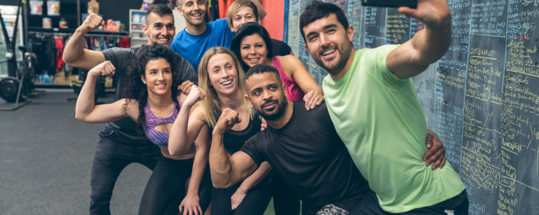 Gruppe von glücklichen Fitness-Enthusiasten posiert für ein Selfie im Fitnessstudio vor einer Tafel mit Trainingsübungen und Notizen.