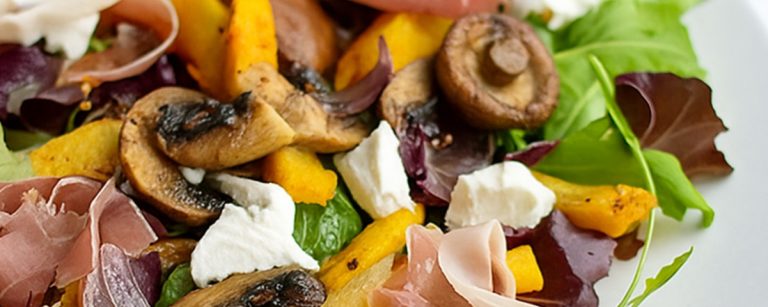 Nahaufnahme einer gesunden und sausgewogenen Mahlzeit, die zarte sScheiben Schinken, cremigen Ziegenkäse, geröstete Pilze und leuchtend orangefarbenen Kürbis über einem Bett aus frischen grünen und roten Blättern präsentiert.