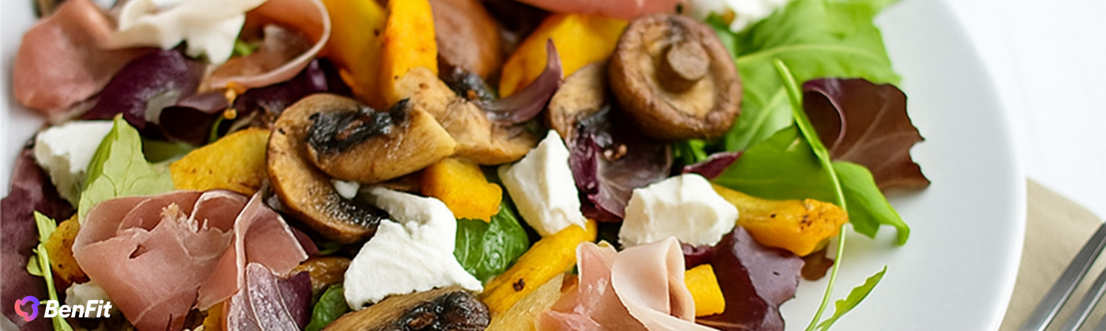 Nahaufnahme einer gesunden und sausgewogenen Mahlzeit, die zarte sScheiben Schinken, cremigen Ziegenkäse, geröstete Pilze und leuchtend orangefarbenen Kürbis über einem Bett aus frischen grünen und roten Blättern präsentiert.