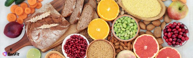 Gevarieerde selectie van gezonde voedingsmiddelen zoals volkorenbrood, vers fruit (sinaasappels, grapefruit, appel, cranberries, granaatappel), knapperige groenten (wortels, komkommer, ui), peulvruchten (linzen, edamame), volkorenproducten (gierst, tarwe) en noten/zaden (amandelen, lijnzaad). Ideaal voor een evenwichtige, vezelrijke en voedingsrijke fitnessvoeding.