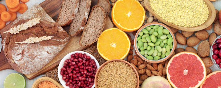 Gevarieerde selectie van gezonde voedingsmiddelen zoals volkorenbrood, vers fruit (sinaasappels, grapefruit, appel, cranberries, granaatappel), knapperige groenten (wortels, komkommer, ui), peulvruchten (linzen, edamame), volkorenproducten (gierst, tarwe) en noten/zaden (amandelen, lijnzaad). Ideaal voor een evenwichtige, vezelrijke en voedingsrijke fitnessvoeding.